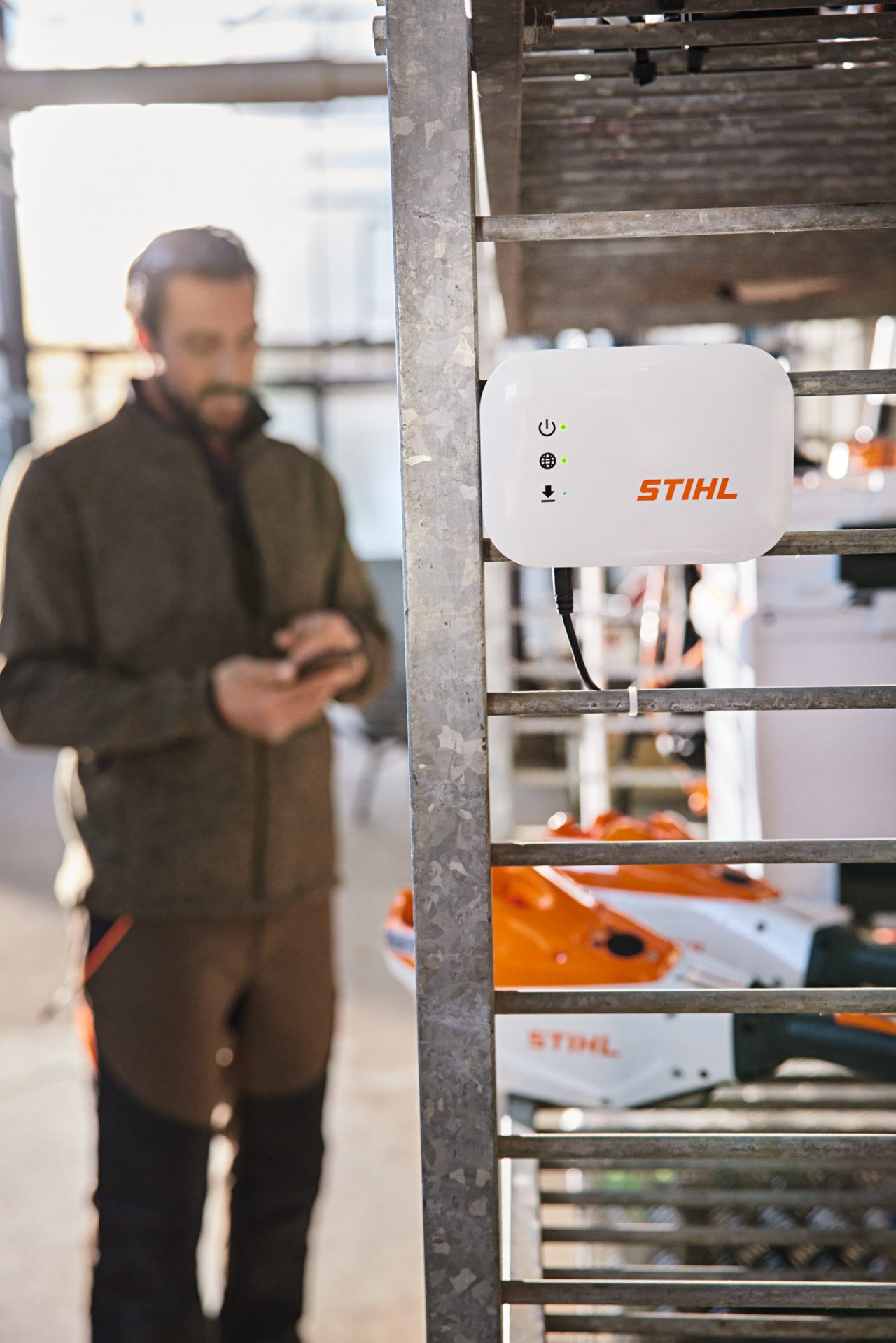 A STIHL connected Box hung on a workshop shelf. In the background there is a gardener and landscaper interacting with their smartphone. 