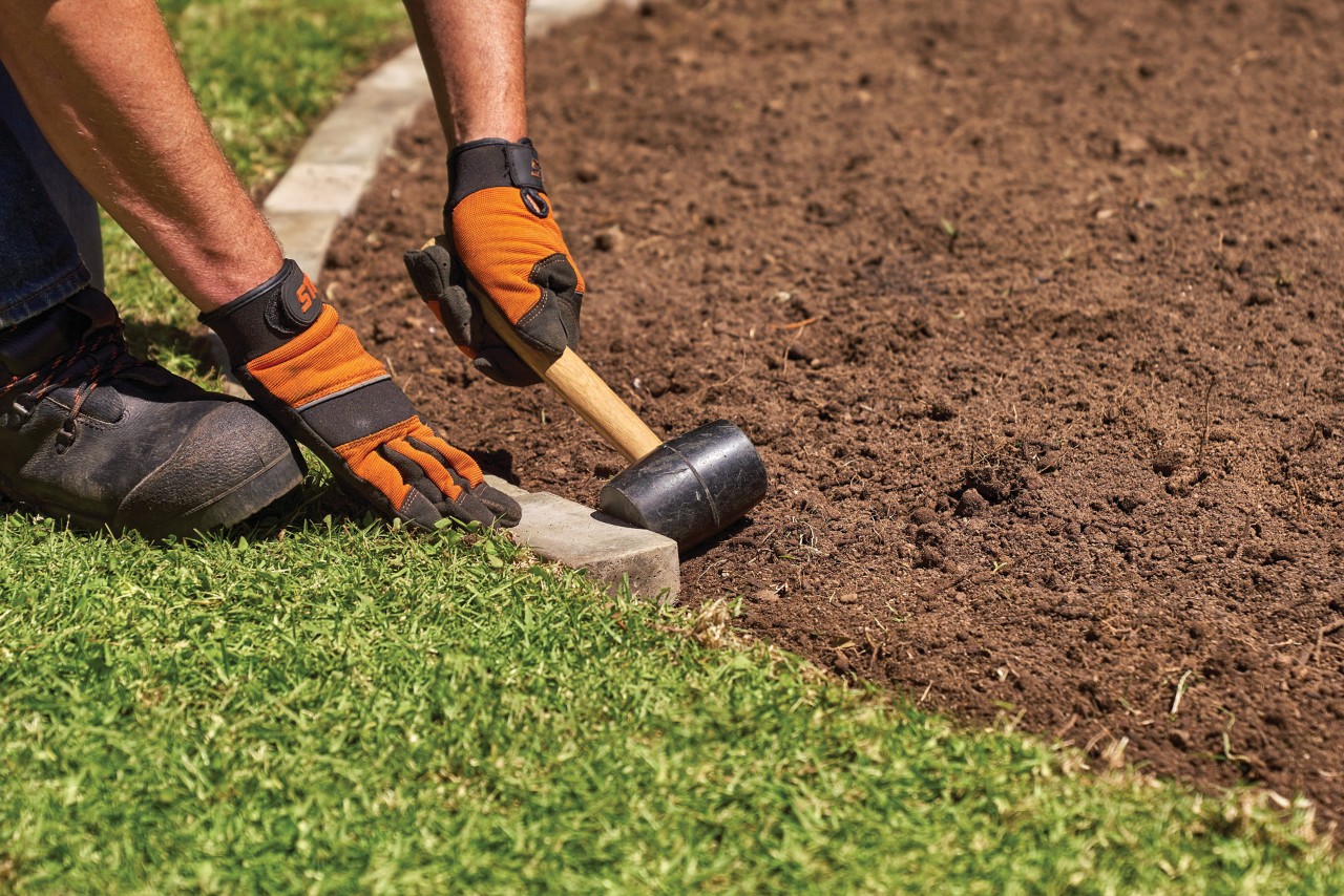 A person wearing gloves using a mallet to create a border of bricks on a garden bed
