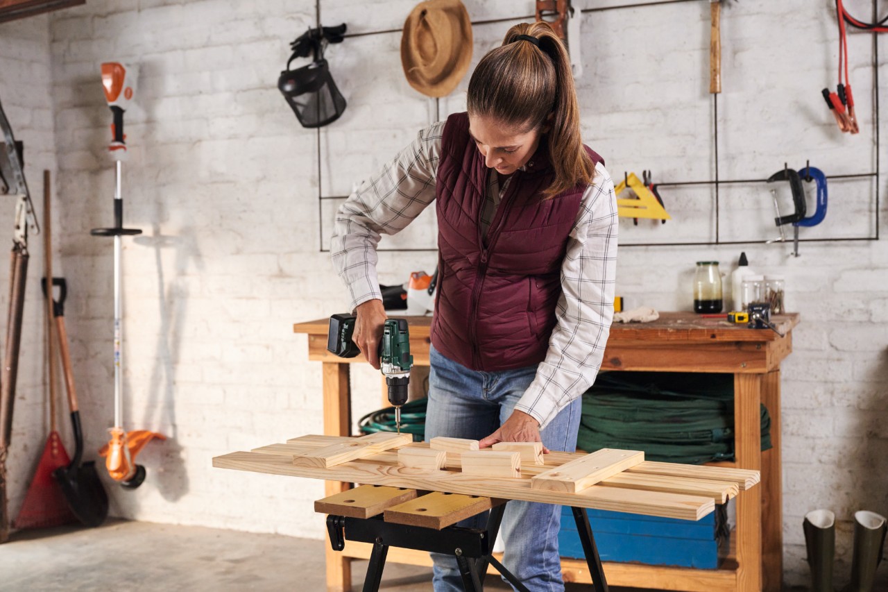 A woman with a checked top and ponytail screwing slats to cross struts on a workbench, with a wall of tools in the background