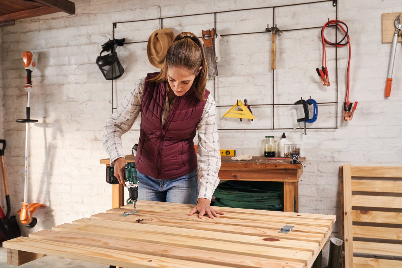 A woman using a cordless screwdriver to screw a removal flap onto the front of the composter on a workbench