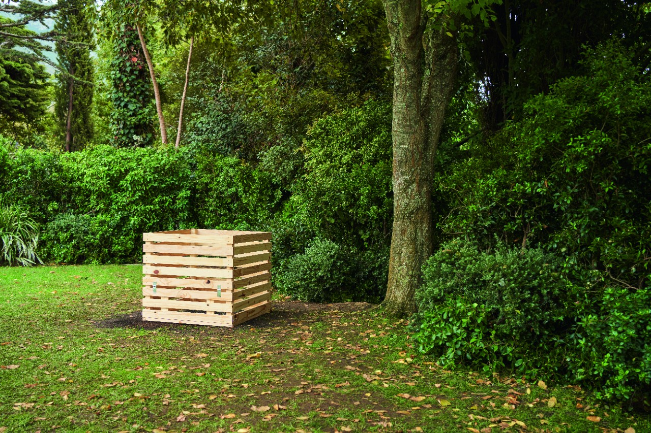 DIY wooden composter on a garden plot, with a dense hedge and trees in the background