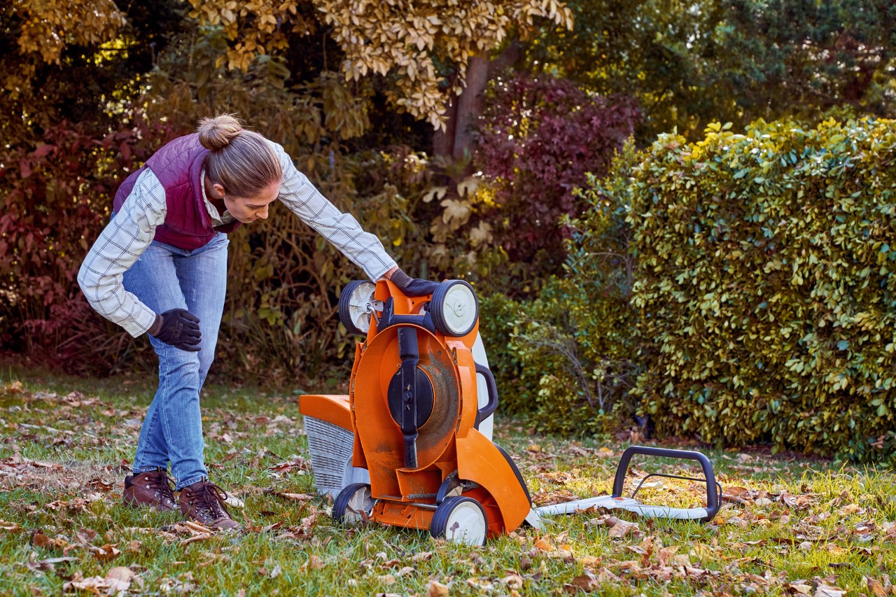 Woman inspecting the underside of a STIHL RMA 339 C lawnmower in a garden during lawn care in autumn