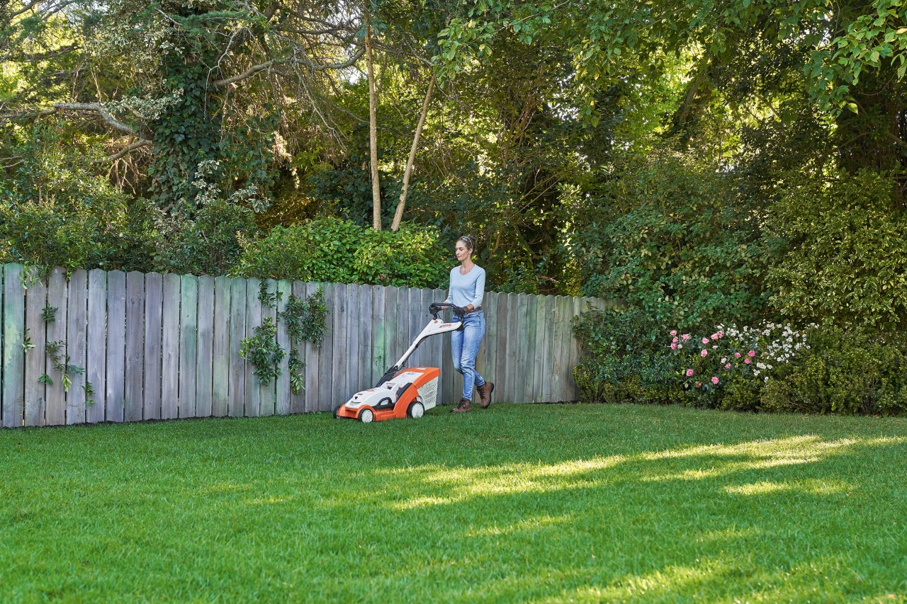 Woman mowing a green lawn in front of a wooden fence with a STIHL RMA 339 C cordless lawn mower.