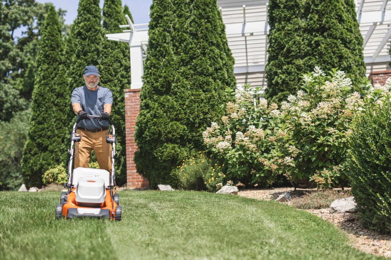 A woman using a STIHL RMA 339 C cordless lawn mower to cut grass with wooden fence and log store in the background