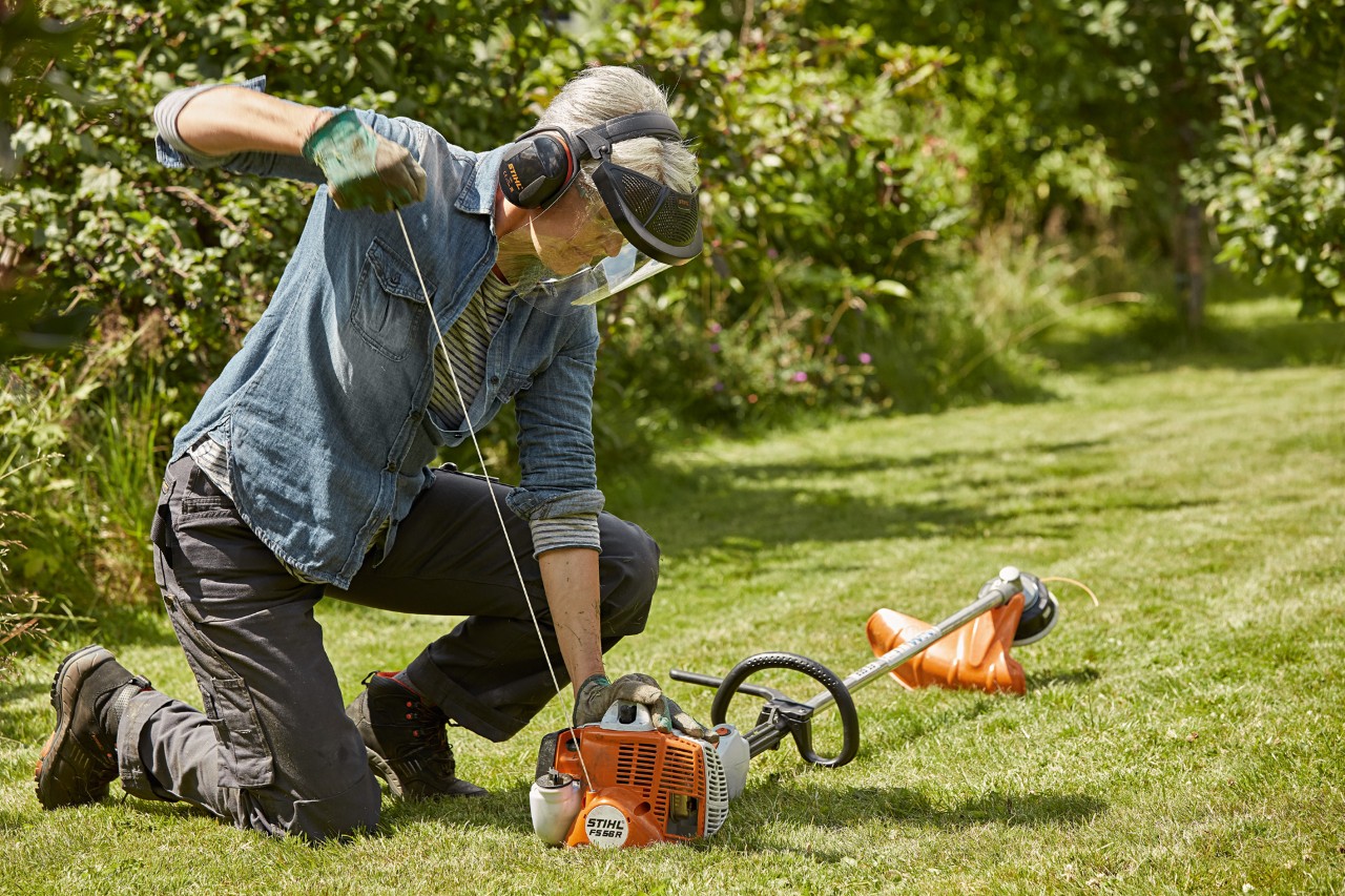 Woman wearing ear and face protection starting a STIHL FS 56 R petrol brushcutter in a grassy area