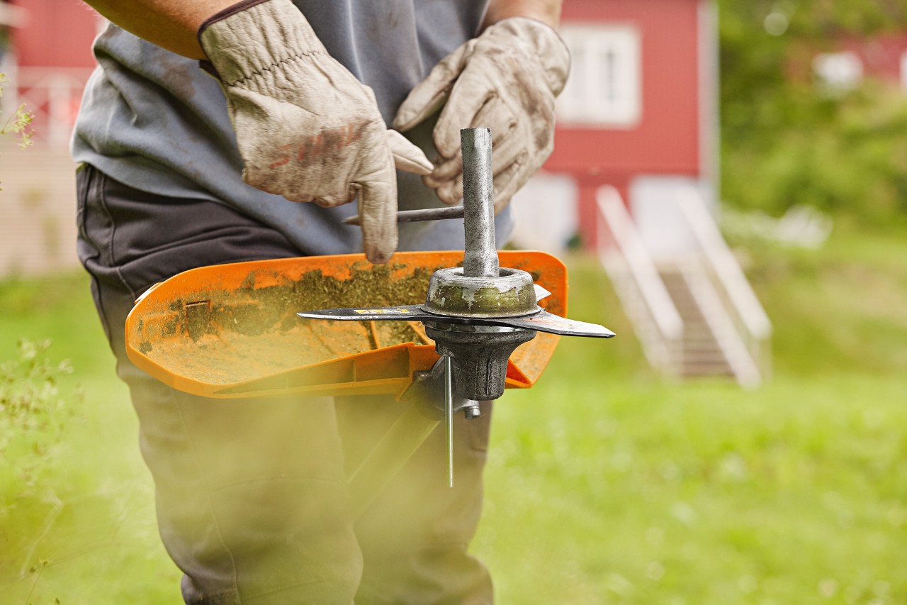 A person in front of a house with green lawn, preparing a brushcutter for a change of cutting tool