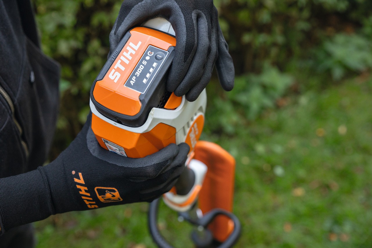 Close-up of someone wearing black protective gloves, holding the STIHL HLA 66 cordless long-reach hedge trimmer and handling the STIHL AP 300 S lithium-ion battery